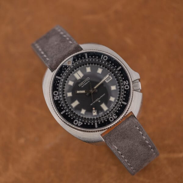 Angled Seiko 6105-8119 diver showing aged hands, faded bezel pip and anthracite suede strap