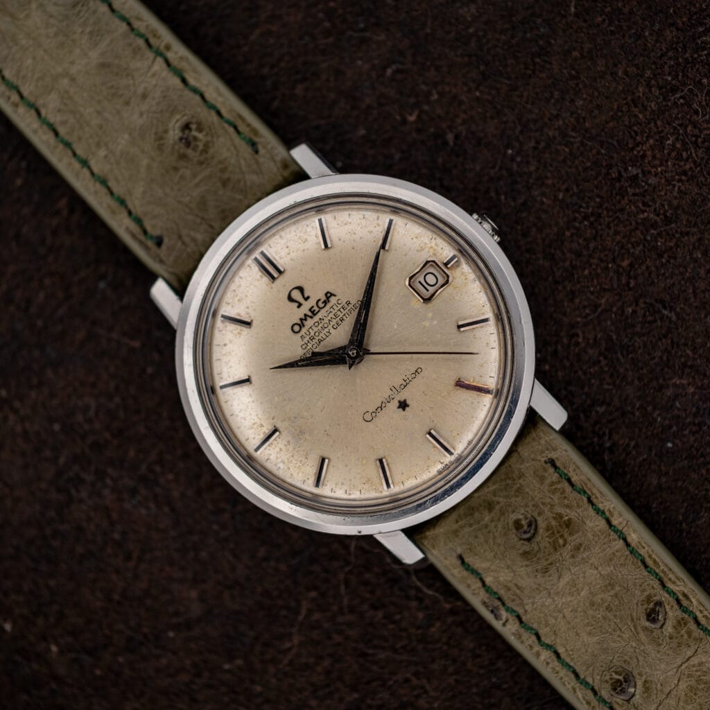 Omega Constellation 168.004 resting diagonally on brown fabric with its silver dial and olive ostrich strap