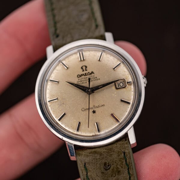 Omega Constellation 168.004 held between fingers, with the silver dial, date window and olive strap visible