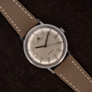 Movado Kingmatic 15161 with silver sunburst dial and taupe leather strap resting diagonally on dark cloth