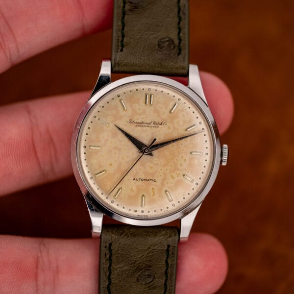 IWC held upright between fingers, displaying its mottled eggshell dial and olive ostrich strap