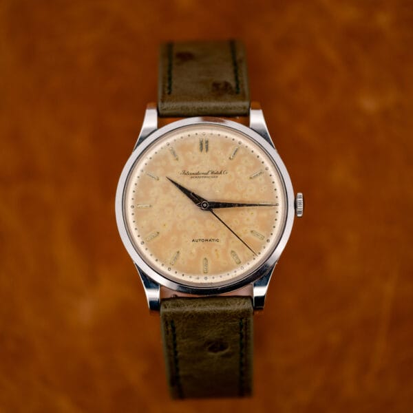 Straight-on IWC view with central seconds, paired twelve o'clock markers and the olive strap curved underneath
