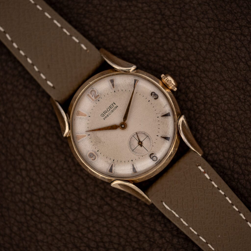 Gruen Precision 415/932 with curved fancy lugs and taupe leather strap resting diagonally on a textured brown surface