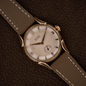 Gruen Precision 415/932 with curved fancy lugs and taupe leather strap resting diagonally on a textured brown surface