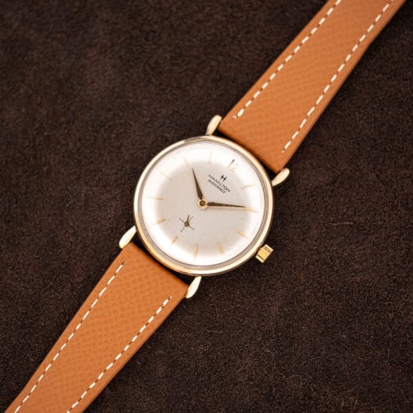 Overhead view of Hamilton Masterpiece with tan leather strap extended across dark brown cloth