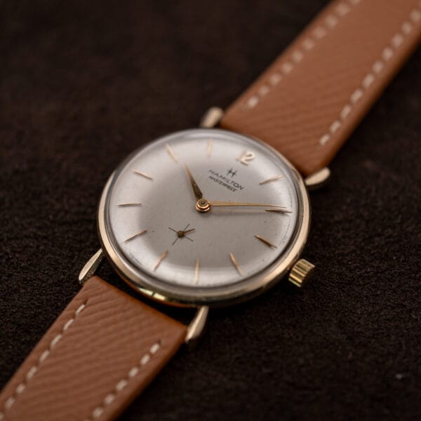 Angled Hamilton Masterpiece showing its raised crystal, winding crown and tan strap extending toward the upper right