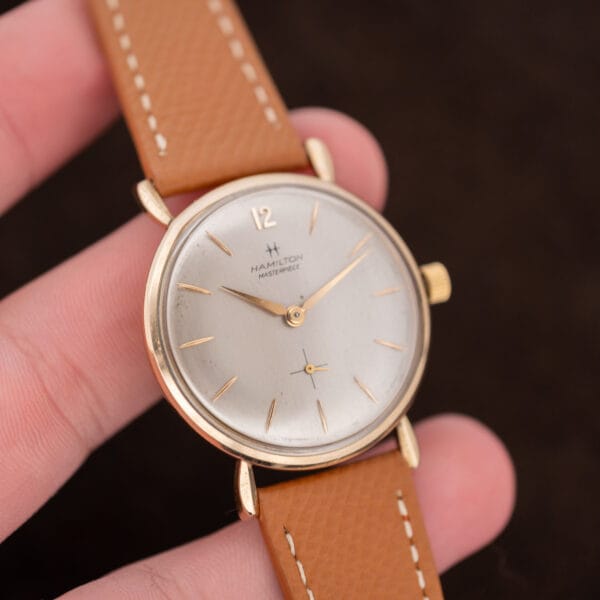 Hamilton Masterpiece held upright between fingers, showing the full silver dial and tan leather strap