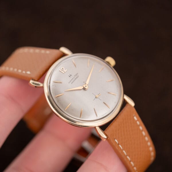 Hand-held Hamilton Masterpiece with its tan strap arched behind the case and lower lugs in view