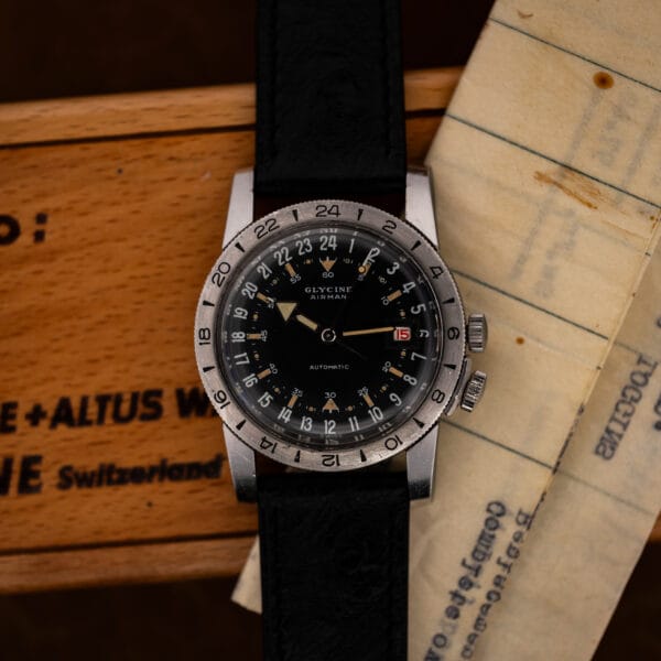Glycine Airman face-up on its wooden box beside folded paperwork, with the black 24-hour dial centered