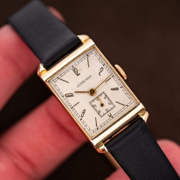 Longines rectangular watch held between fingers, displaying the off-white dial and slender Arabic numerals