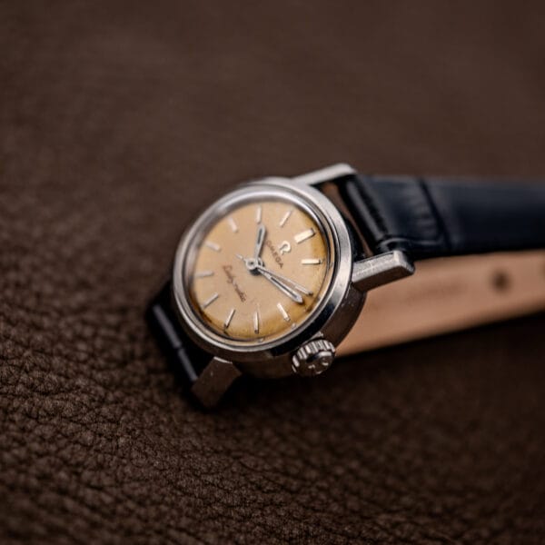 Omega Ladymatic 11000 SC resting at an angle with curved black strap and visible tan lining