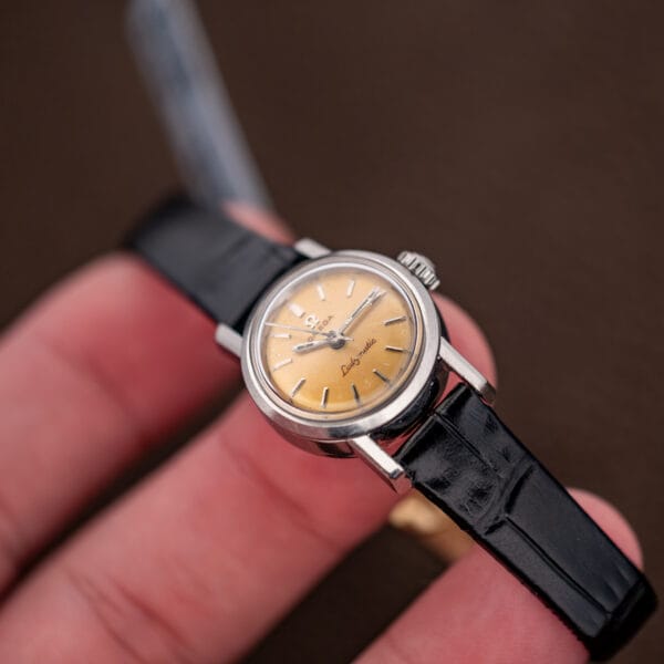 In-hand oblique view of Omega Ladymatic with lemon dial, smooth bezel and black patterned strap