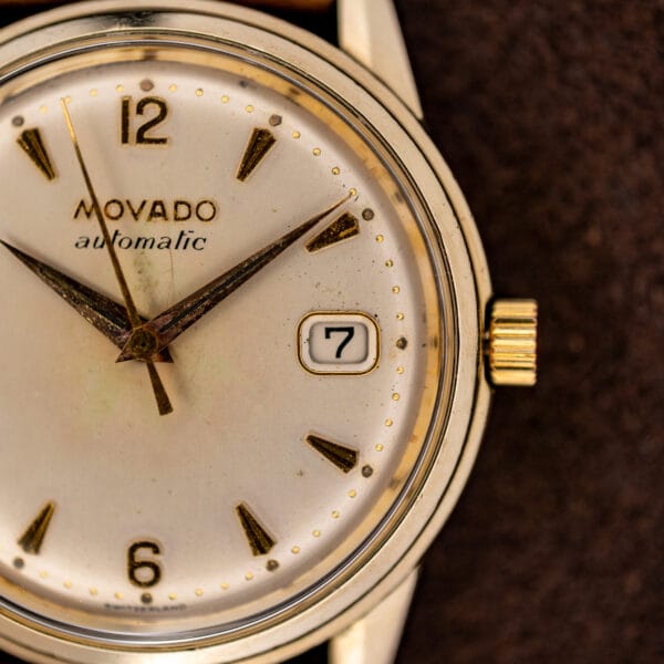 Movado Automatic date aperture close-up with rounded gold-tone frame beside the winding crown