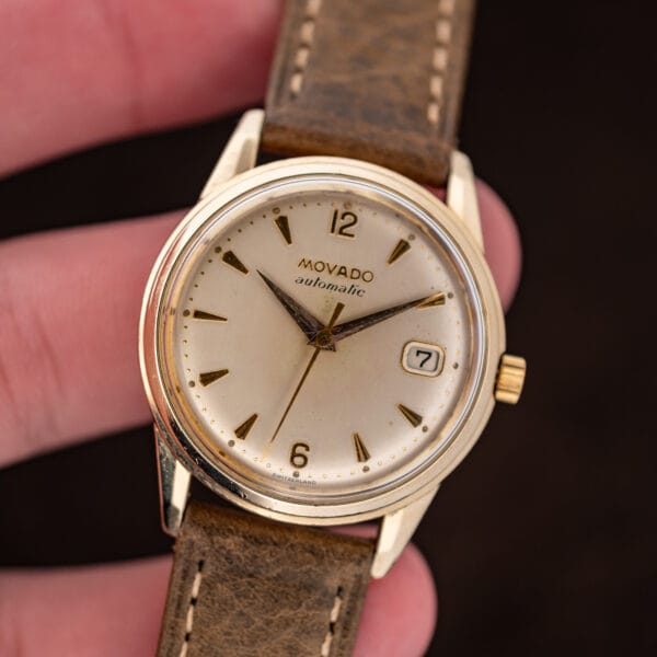 Hand-held Movado 64389 showing white dial, Arabic numerals and framed date display