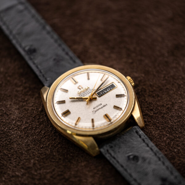 Omega Seamaster low diagonal view with day-date display, angular lugs and textured black strap against brown background
