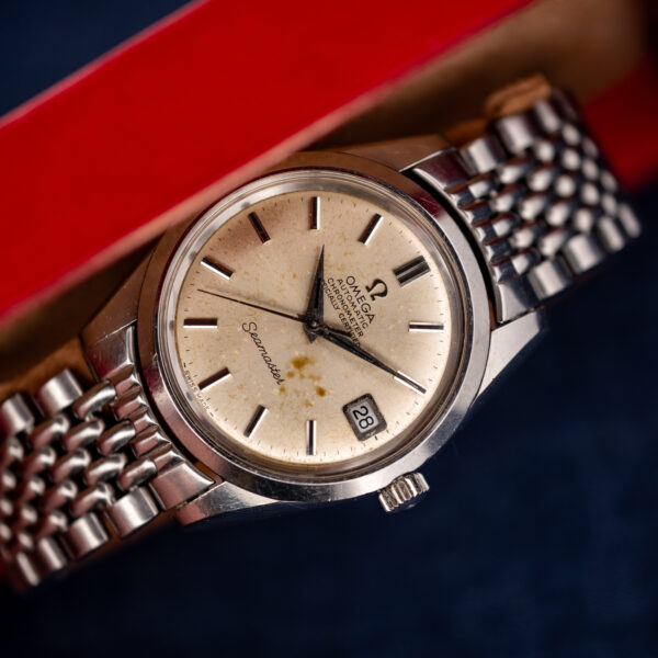 Omega Seamaster chronometer beside red tray edge, showing date dial and stainless steel bracelet on blue fabric