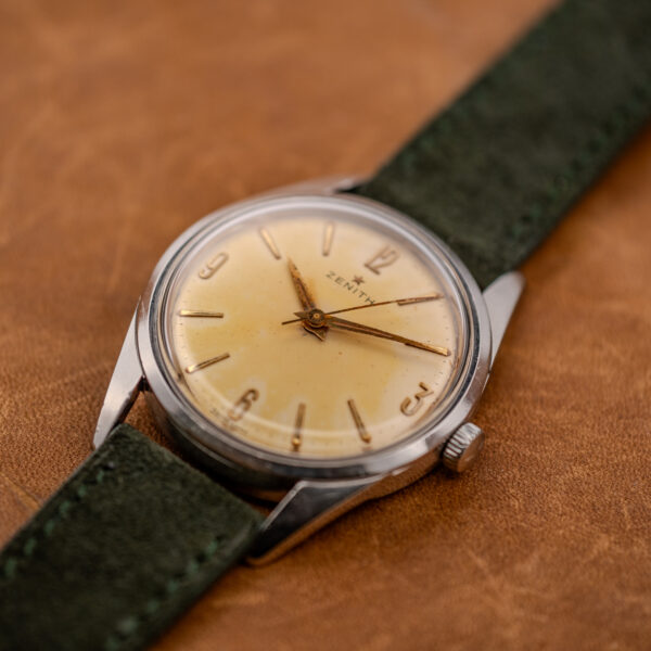 Zenith crown-side view with Arabic numeral dial, pointed hands and raised crystal beside green suede strap