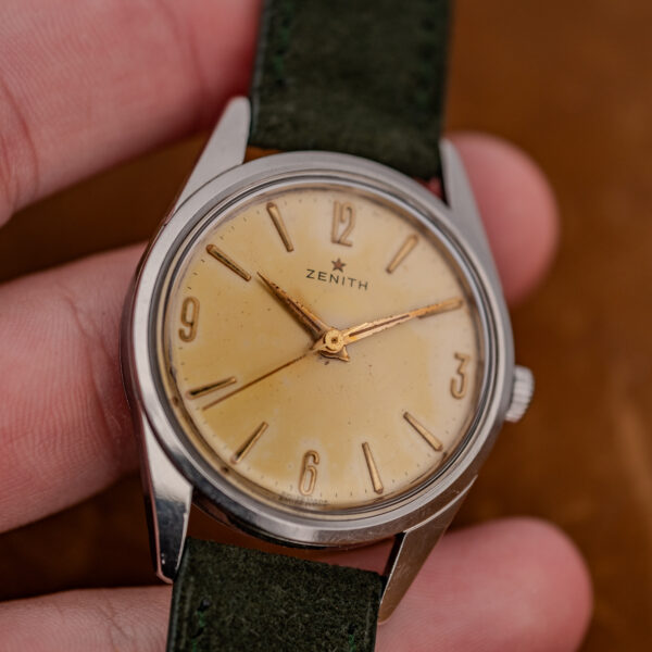 Zenith manual wind watch held in hand, showing Arabic quarter-hour numerals and forest green suede strap