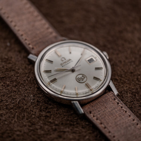 Omega automatic opposite diagonal view showing USS emblem, baton markers and stitched strap beside the case