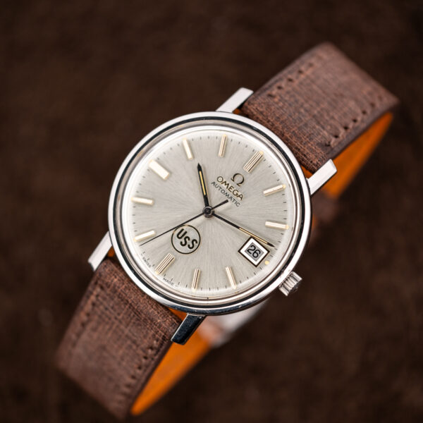 Omega automatic upright portrait with silver sunburst dial, circular USS emblem and stitched strap looped beneath