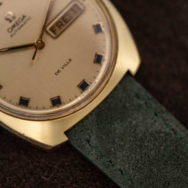 Corner of the gold-plated Omega De Ville cushion case beside the De Ville dial text and crystal edge