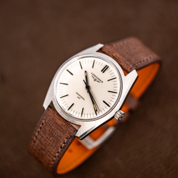 Longines Admiral HF face and brown crosshatched strap raised toward the camera, steel shoulders in view