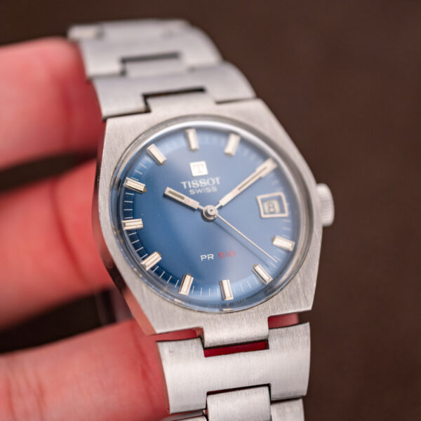 Handheld Tissot PR 516 at an angle, showing the blue dial and wide steel H-link bracelet