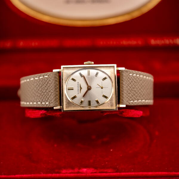 Longines gold-filled tank centered in a red box with the taupe strap spread across the cushion