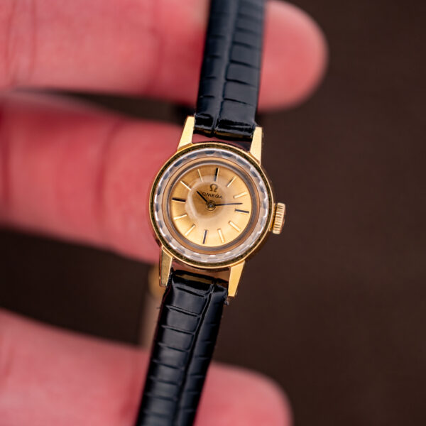 Omega 511.166 held in hand, faceted crystal and gold-tone case beside the black strap